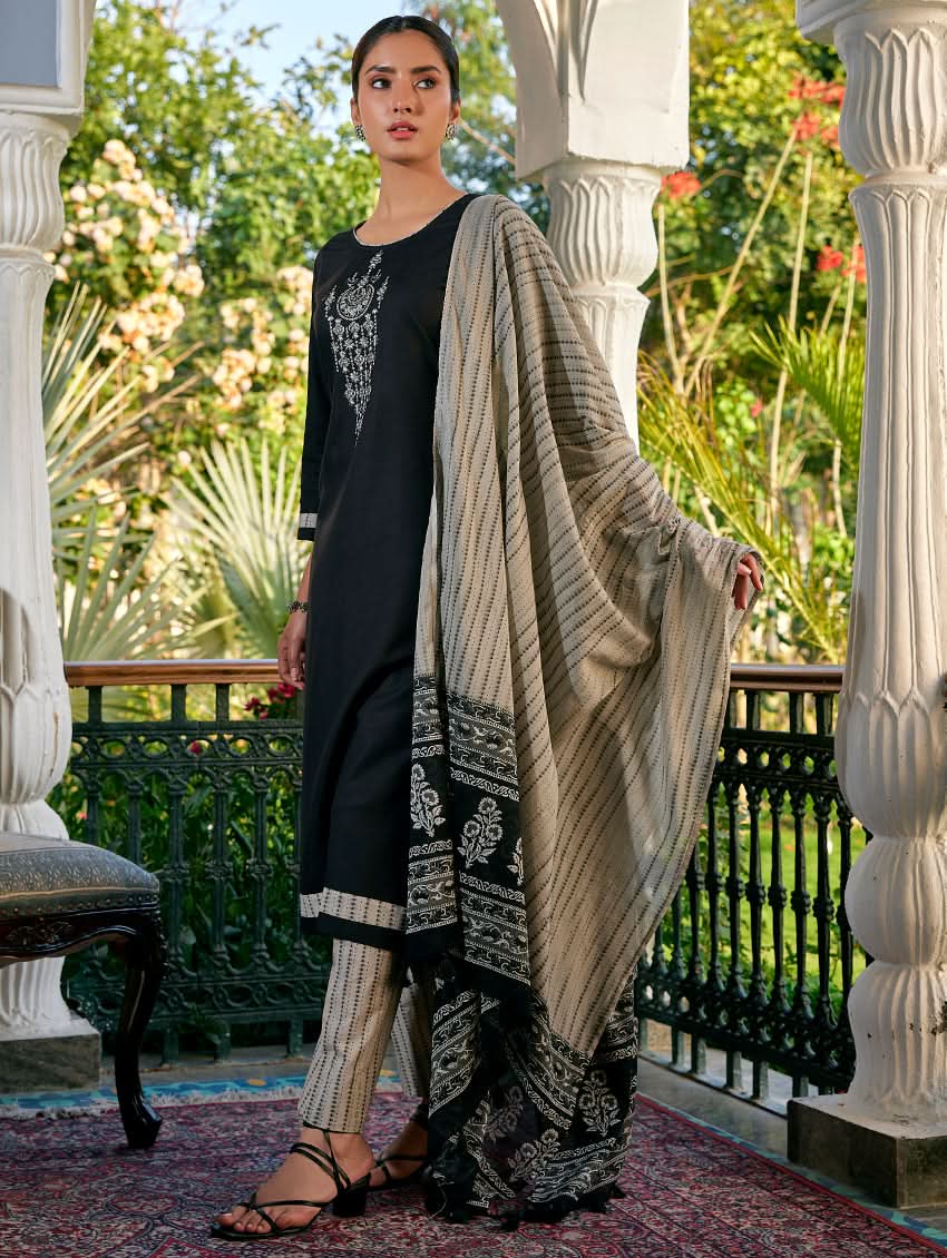 A woman standing in a columned hallway wearing a Black silk suit set with embroidered details, consisting of a kurta with round neckline and three-fourth sleeves, and pants with a partially elasticated waistband. The dupatta is also visible and printed,