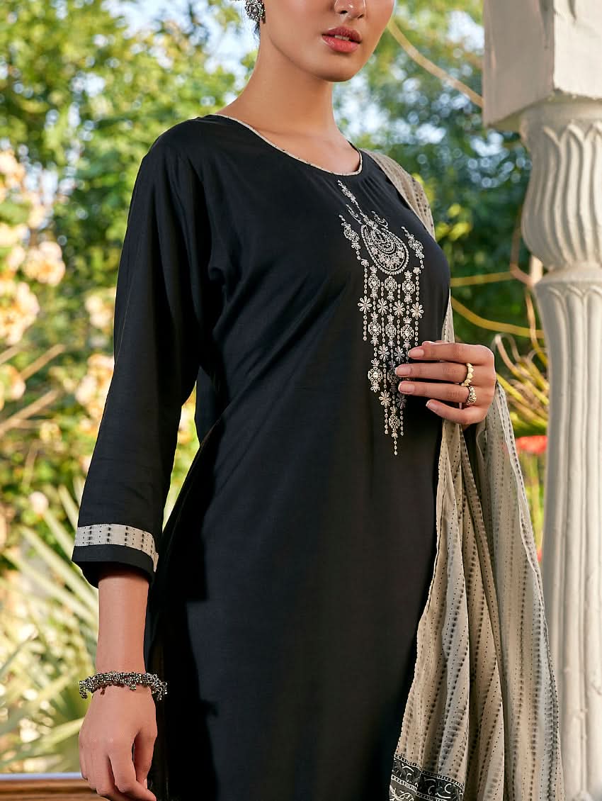 A woman standing in a columned hallway wearing a Black silk suit set with embroidered details, consisting of a kurta with round neckline and three-fourth sleeves, and pants with a partially elasticated waistband. The dupatta is also visible and printed,