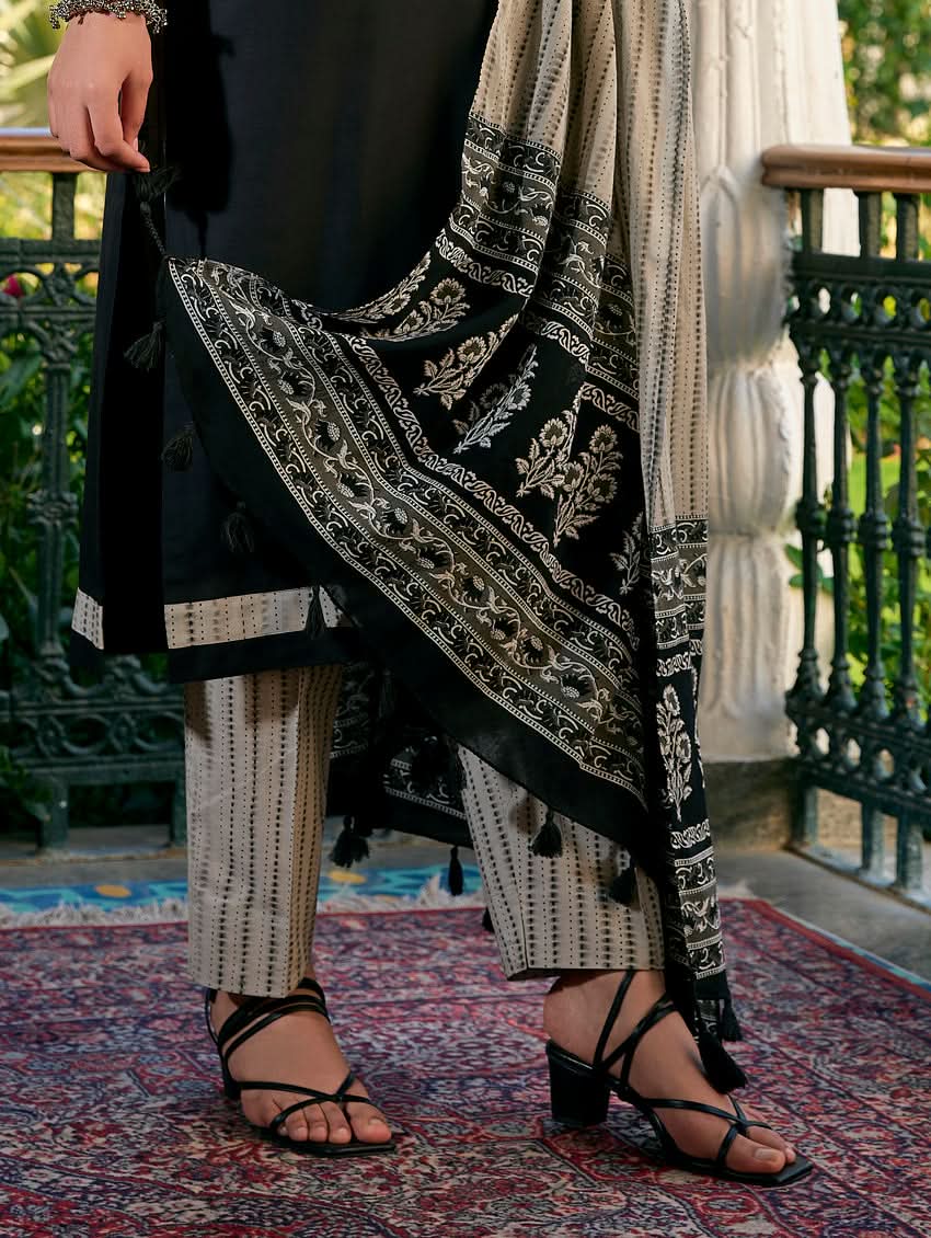 A woman standing in a columned hallway wearing a Black silk suit set with embroidered details, consisting of a kurta with round neckline and three-fourth sleeves, and pants with a partially elasticated waistband. The dupatta is also visible and printed,