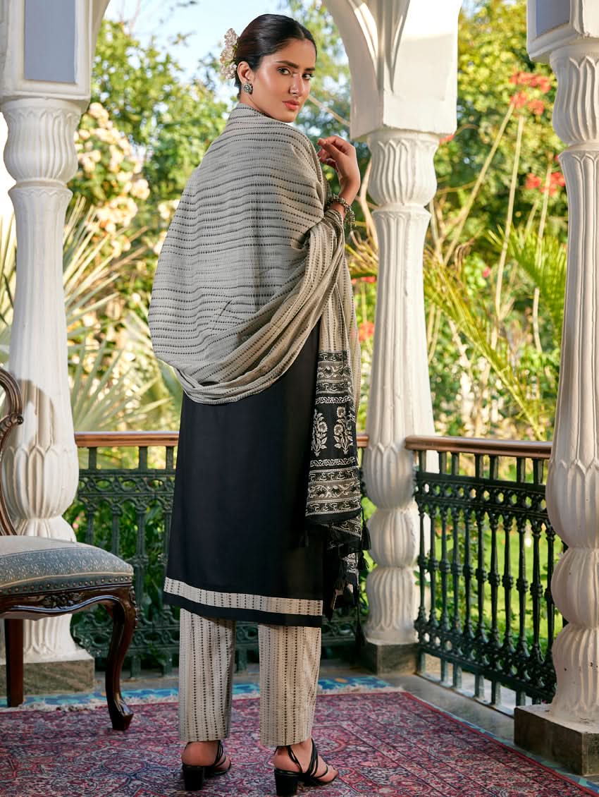 A woman standing in a columned hallway wearing a Black silk suit set with embroidered details, consisting of a kurta with round neckline and three-fourth sleeves, and pants with a partially elasticated waistband. The dupatta is also visible and printed,