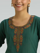 Woman wearing a green top with intricate embroidery on a plain background