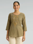 Woman wearing an olive green top with gold embroidery on a gray background