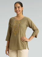 Woman wearing an olive green top with gold embroidery on a plain background