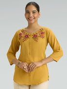 Woman wearing a yellow embroidered top against a plain background