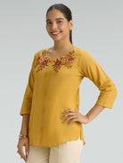 Woman wearing a mustard yellow top with floral embroidery on a gray background