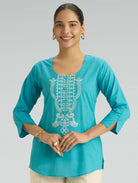 Woman wearing a turquoise embroidered top on a gray background
