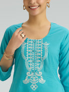 Woman wearing a turquoise top with white embroidery on a plain background