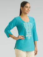 Woman wearing a turquoise top with white embroidery on a gray background