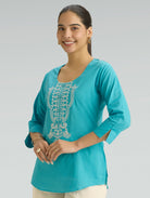 Woman wearing a turquoise top with intricate white embroidery on a gray background
