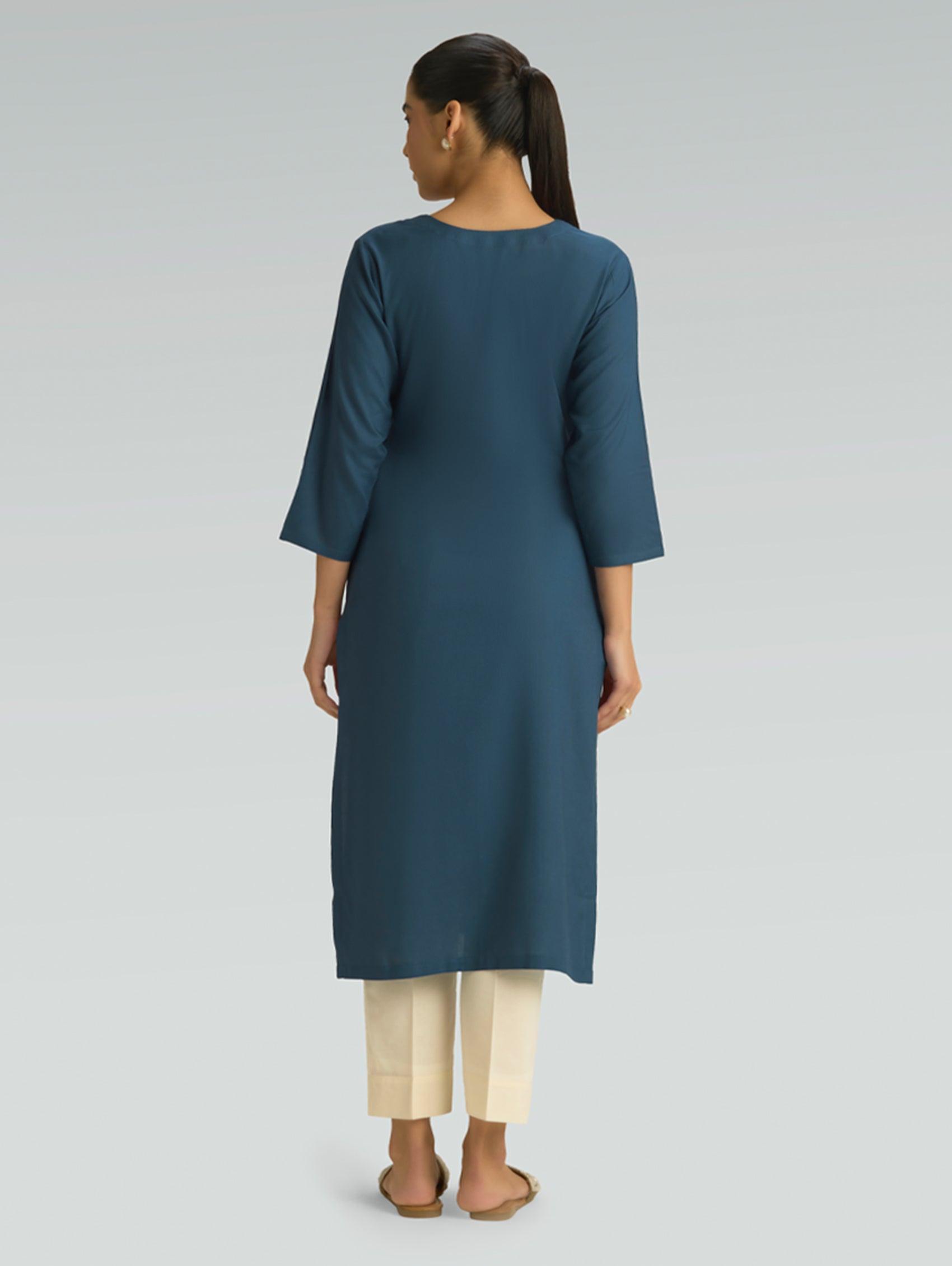 “A woman dressed in a teal kurta paired with beige pants, captured in a graceful back pose on a light gray background.”