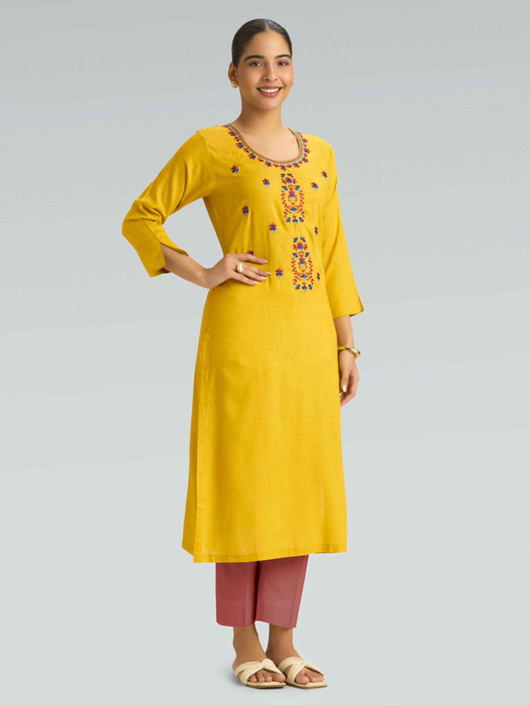 “A woman wearing a straight-fit yellow kurta with a round neckline and computerized thread embroidery, paired with pink pants, set against a gray background.”