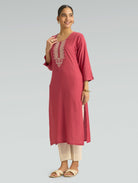 A woman wearing a straight-fit pink kurta with a round neckline, adorned with computerized thread embroidery, set against a soft gray background.