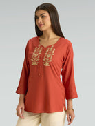Woman wearing a rust-colored top with gold embroidery on a gray background