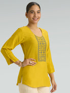 Woman wearing a yellow top with intricate embroidery on a gray background