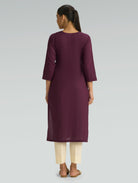 A woman in a purple kurta paired with beige pants, shown in a back pose against a light gray background.