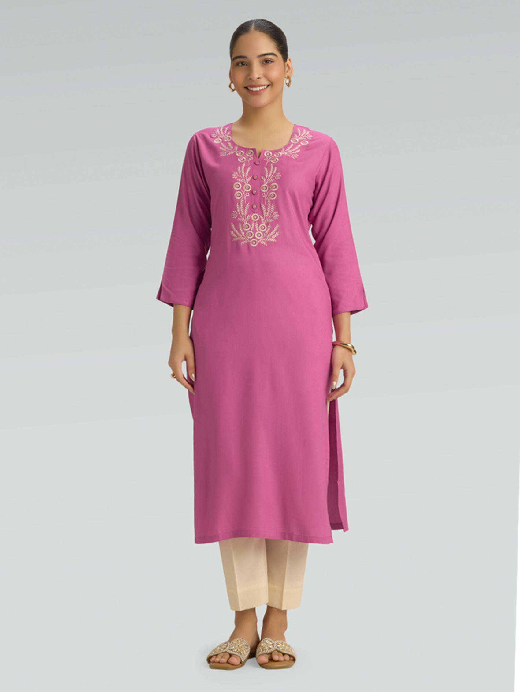 Woman wearing a pink embroidered kurta with a round neckline and 3/4 sleeves, paired with beige pants on a gray background.