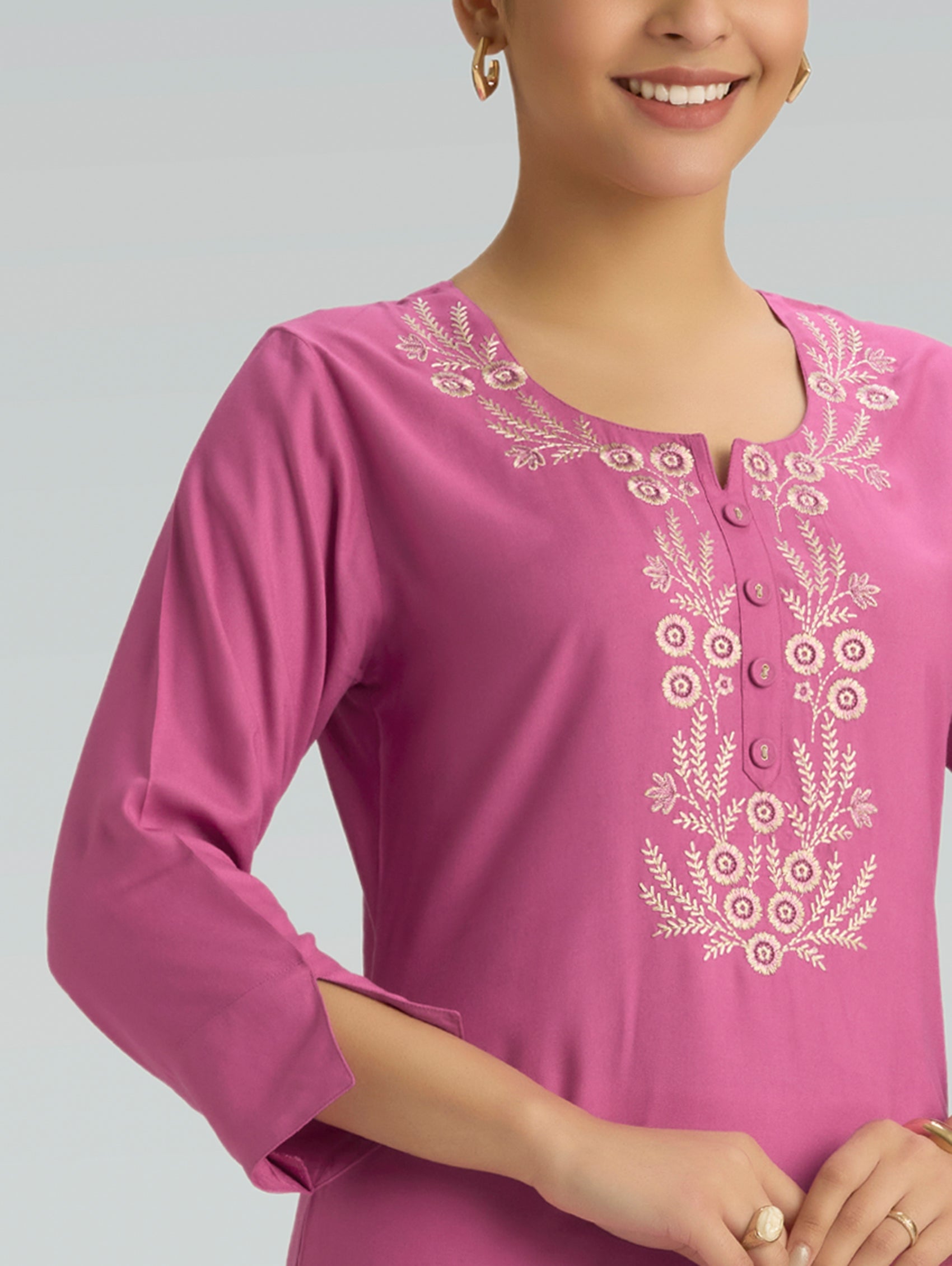 A woman wearing a pink top with white embroidery along with 3/4 sleeves on a grey background