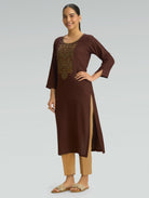 A woman is wearing a brown kurta with a side slit and gold embroidery, along with 3/4 sleeves, on a gray background.