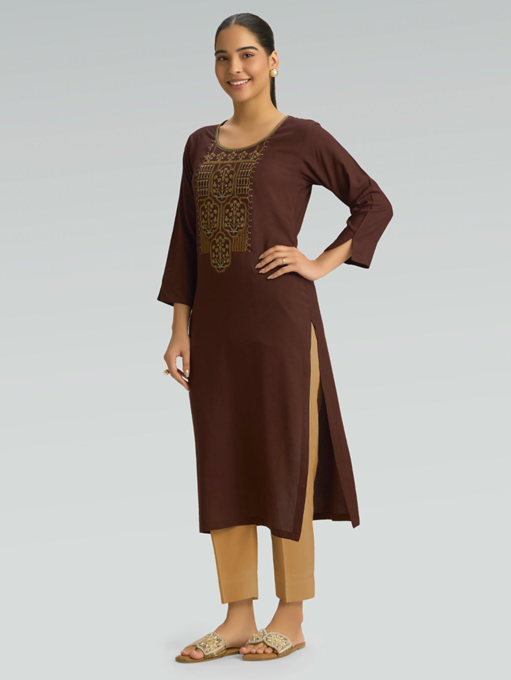 A woman is wearing a brown kurta with a side slit and gold embroidery, along with 3/4 sleeves, on a gray background.