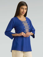 Woman wearing a blue top with intricate embroidery on a gray background