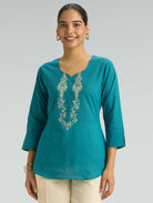 Woman wearing a teal top with intricate silver embroidery on a plain background