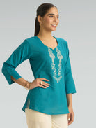 Woman wearing a teal top with floral embroidery on a gray background