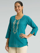 Woman wearing a teal top with intricate silver embroidery on a gray background