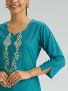 Woman wearing a teal dress with intricate white embroidery on a gray background