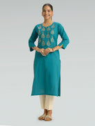 A woman wearing an Iris Blue Colour kurta with computer thread embroidered and a round neckline with 3/4 Sleeves with a side Slit and  beige pants on a grey background