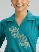 Teal shirt with floral embroidery worn by a person on a gray background