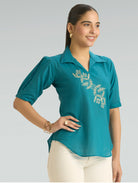 Woman wearing a teal blouse with floral embroidery on a gray background