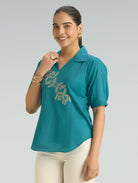 Woman wearing a teal blouse with floral embroidery on a gray background