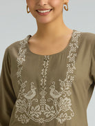 Woman wearing a brown top with intricate white embroidery on a plain background