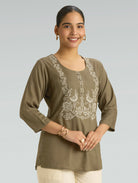 Woman wearing an olive green embroidered top against a plain background