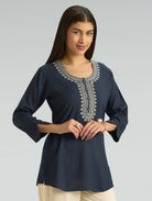 Woman wearing a navy blue top with intricate white embroidery on a gray background
