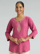 Woman wearing a pink embroidered top against a gray background