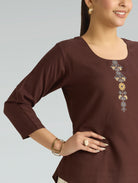 Woman wearing a brown long-sleeve top with floral embroidery on a gray background