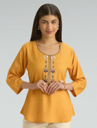 Woman wearing a mustard yellow top with intricate detailing on a gray background