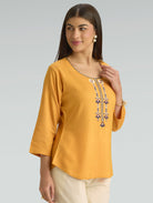 Woman wearing a mustard yellow top with intricate designs on a gray background