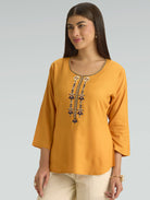 Woman wearing a mustard yellow top with intricate beadwork on a gray background