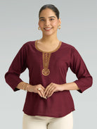 Woman wearing a burgundy top with gold embroidery on a gray background