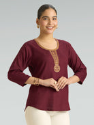 Woman wearing a maroon top with gold embroidery on a gray background