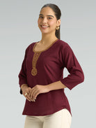 Woman wearing a burgundy top with gold embroidery on a gray background