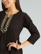 Woman wearing a black long-sleeve top with gold embroidery on a gray background