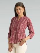 Woman wearing a maroon patterned blouse with white pants on a gray background