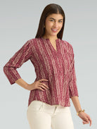 Woman wearing a patterned maroon top with white pants on a gray background